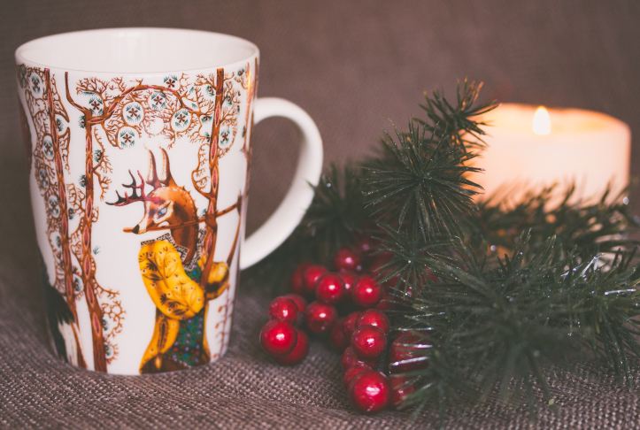 white and gold mug with stag playing flute, in background is a candle, ferns and red berries